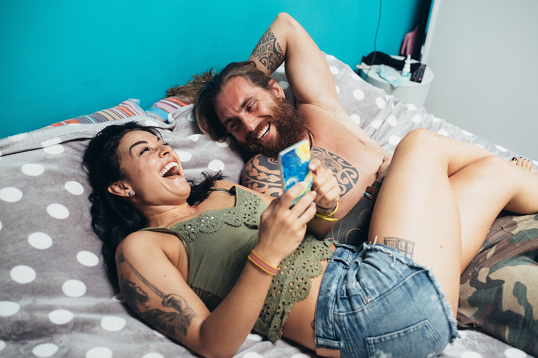 Bearded tattooed man with long brunette hair and woman with long brown hair lying on a bed, laughing.