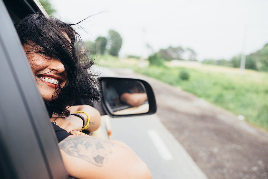 Smiling woman with long brown hair and tattoos looking out of car window.