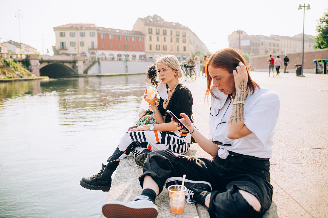 Drei junge Frauen sitzen mit Drinks und Handy an einem Flussufer