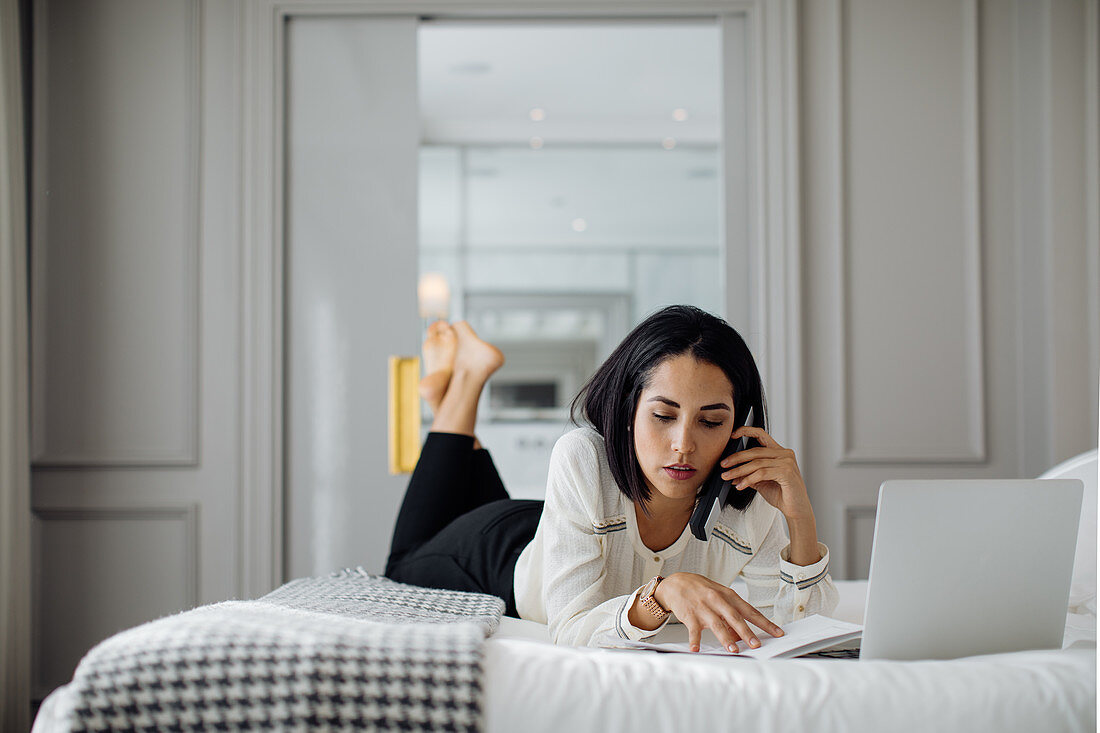 Geschäftsfrau mit Smartphone und Laptop auf dem Bett in einer Suite