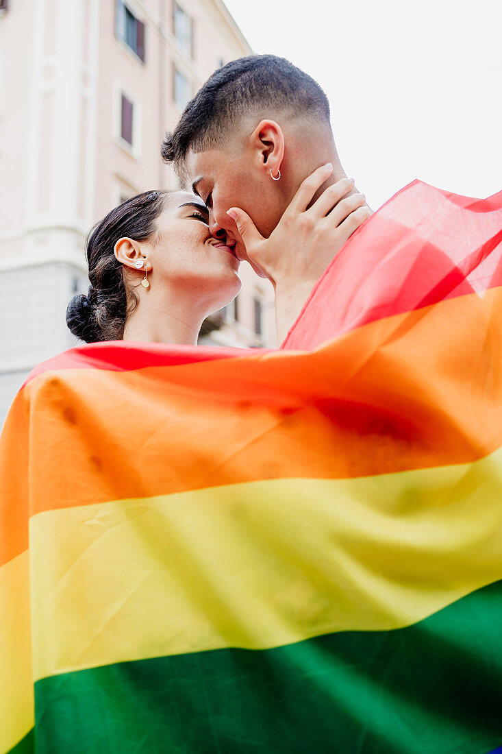 Portrait of young lesbian couple standing on a street, wrapped in rainbow flag, kissing.