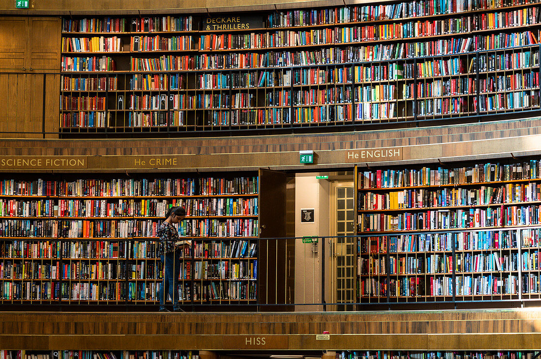 Frau, die ein Buch liest, das neben den Sammlungen auf Regalen, Stadsbiblioteket, öffentliche Bibliothek Stockholm, Schweden steht