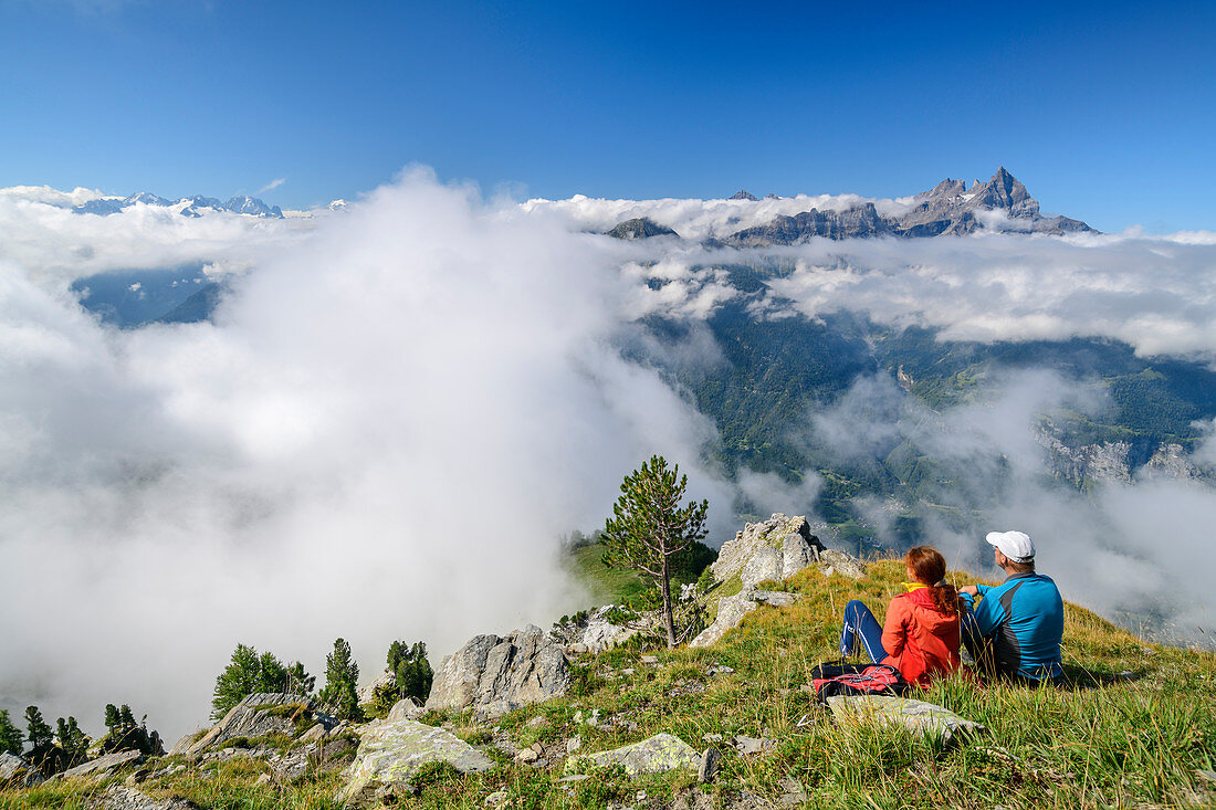 Man and woman while hiking sit on mountain meadow and look at sea of clouds with Dents du Midi, from Dent de Morcles, Bernese Alps, Vaud, Vaud, Switzerland