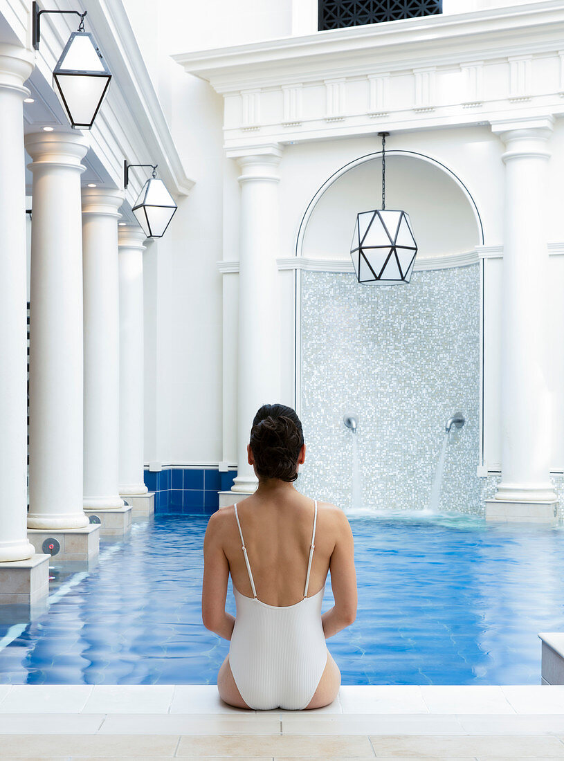 Woman in white bathing suit, sitting with her back to the camera, on the edge of an interior pool. Bath, United Kingdom