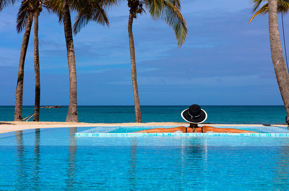 Eine Frau in einem schwarzen Hut mit weißem Rand und dem Rücken zur Kamera ruht auf der Seite eines Pools mit Meerblick. Antigua, Westindische Inseln.
