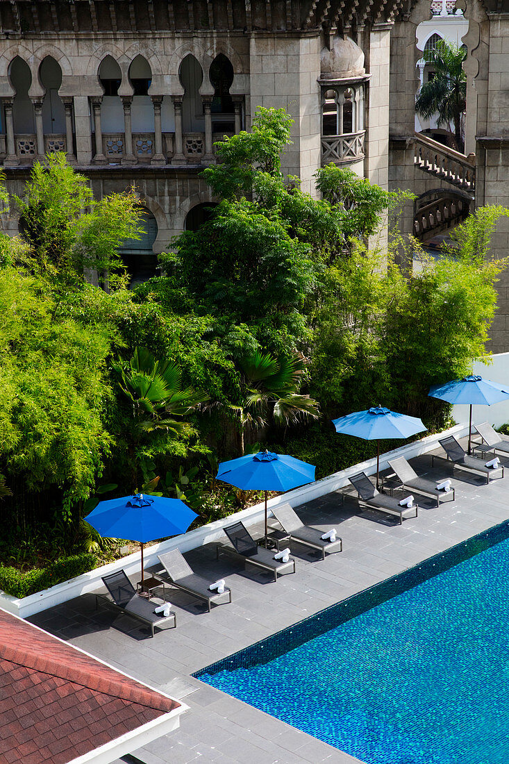Pool with blue umbrellas, set against an old building and lush greenery. Kuala Lumpur, Malaysia.