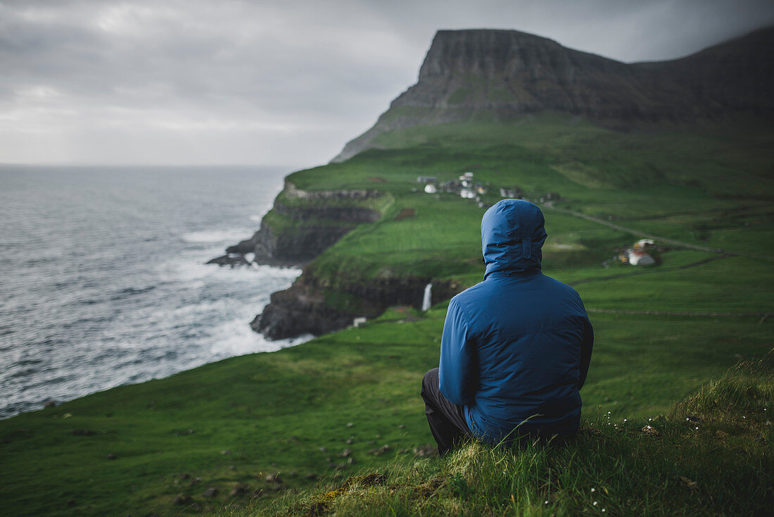 Denmark, Faroe Islands, Gasadalur village, Man sitting and looking at cliffed coast and ocean