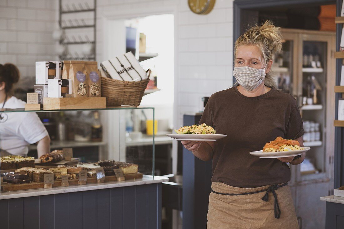 Blond waitress wearing face mask working in a cafe, carrying plates of food.