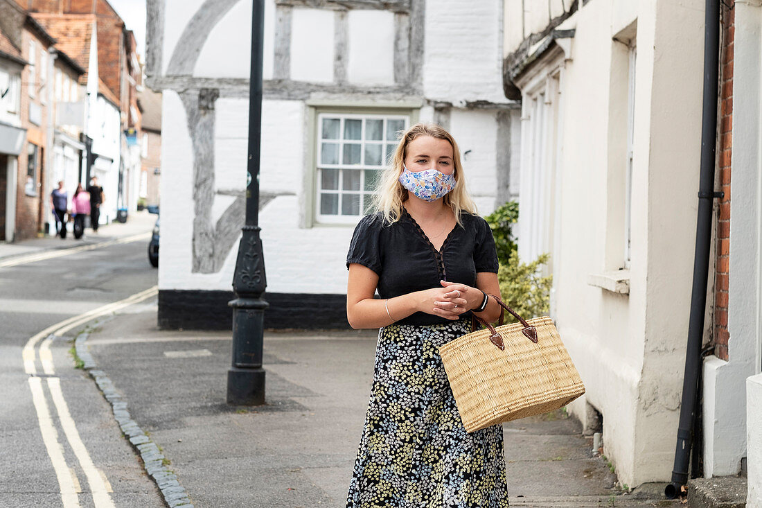 Young blond woman wearing face mask walking through village, carrying shopping bag.