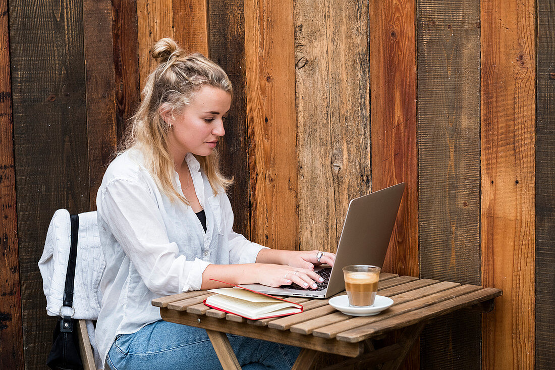 Young blond woman wearing face mask sitting alone at a cafe table with a laptop computer, working remotely.