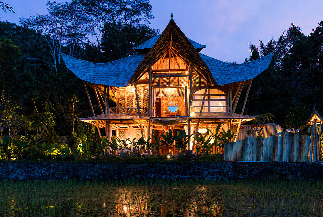 Abendaufnahme eines beleuchteten Bambushauses im Pagodenstil in einem Reisfeld auf Bali, Indonesien, Asien