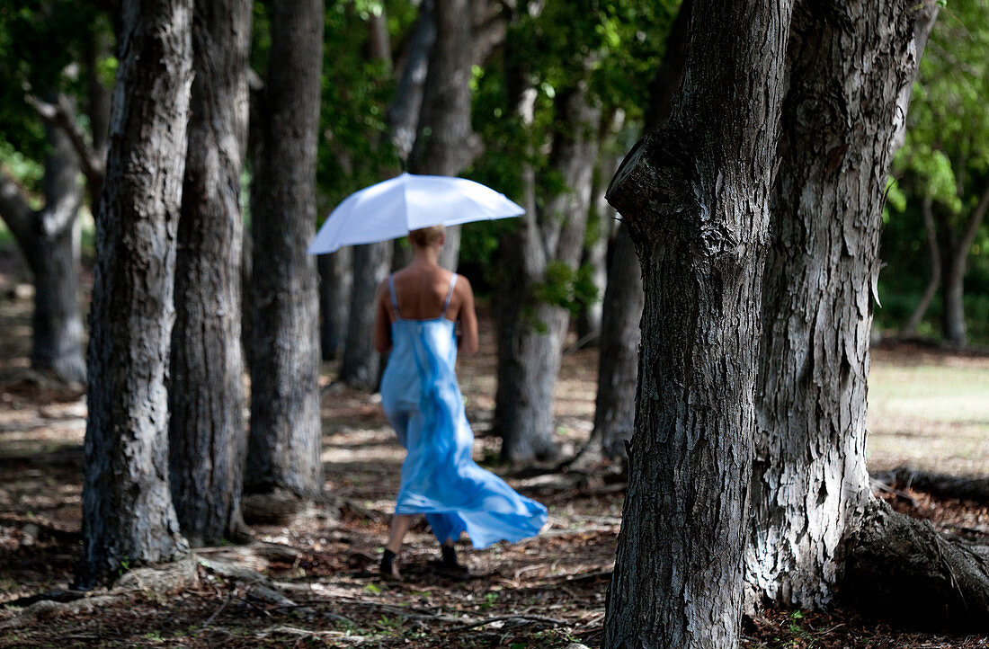 Woman in a blue dress swirling a white umbrella whilewalking in the woods.