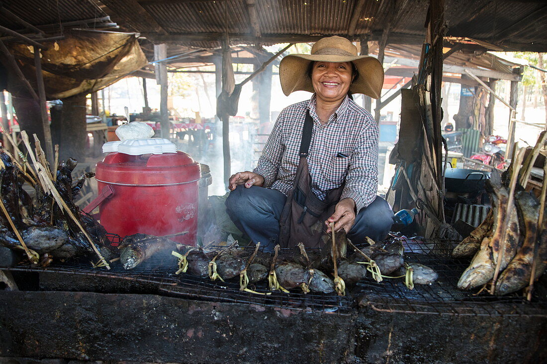 Fröhliche Frau mit gegrilltem Fisch an einem Street Food Verkaufsstand am Markt, Oudong (Udong), Kampong Speu, Kambodscha, Asien