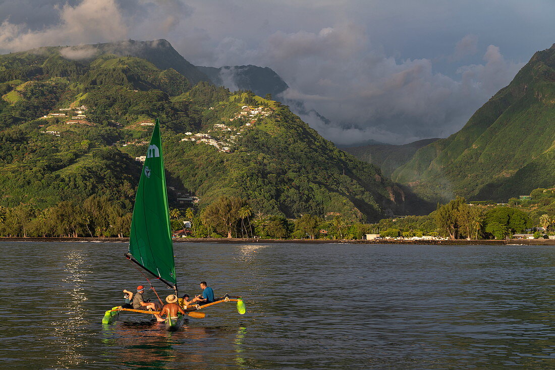 People enjoy a trip on an outrigger canoe with sails in front of a lush mountain backdrop, near Papeete, Tahiti, Windward Islands, French Polynesia, South Pacific