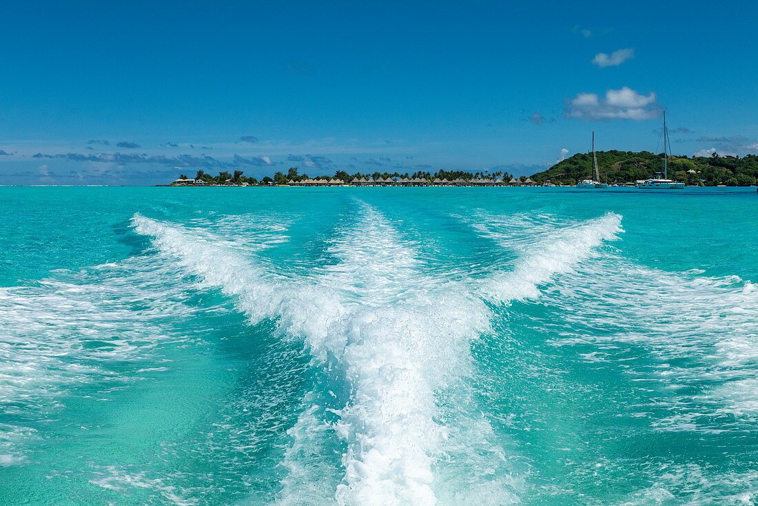 Die Wellen eines Schnellboots im türkisfarbenen Wasser der Lagune von Bora Bora mit Überwasserbungalows in der Ferne, Bora Bora, Leeward Islands, Französisch-Polynesien, Südpazifik