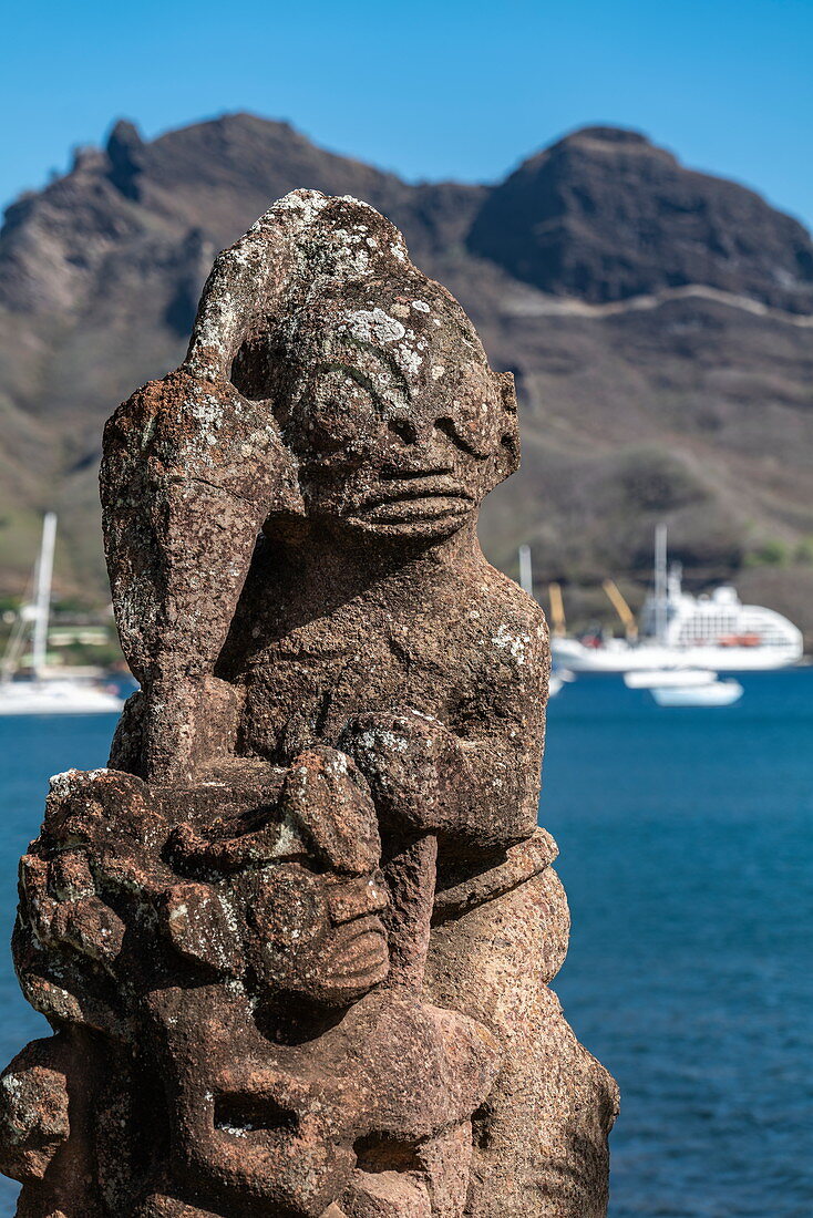 Detail einer Tiki Statue mit Passagierfrachtschiff Aranui 5 (Aranui Cruises) am Pier in der Ferne, Taiohae, Nuku Hiva, Marquesas-Inseln, Französisch-Polynesien, Südpazifik