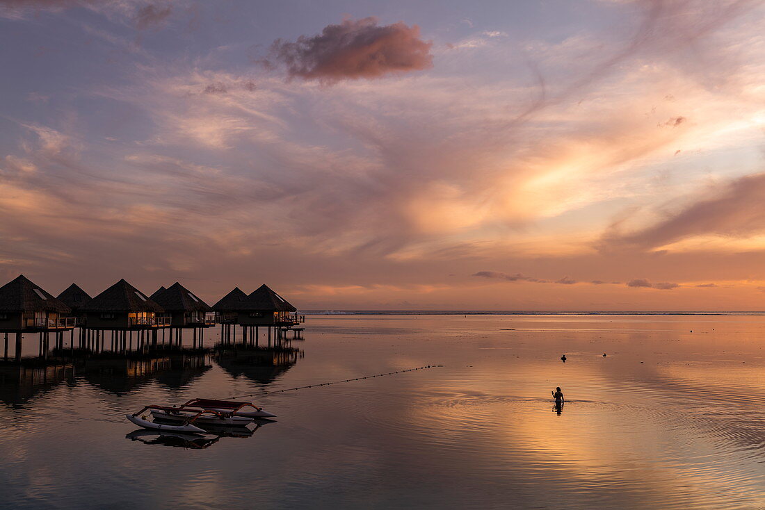 Silhouette von Frau im Wasser neben Auslegerkanu und Überwasserbungalows des Tahiti Ia Ora Beach Resort (managed by Sofitel) bei Sonnenuntergang, nahe Papeete, Tahiti, Windward Islands, Französisch-Polynesien, Südpazifik