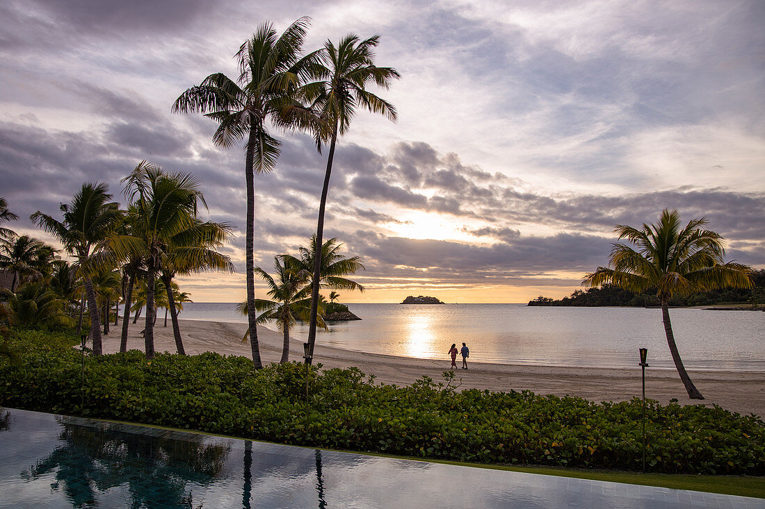 Swimming pool, coconut trees and a couple walking on the beach at Six Senses Fiji Resort at sunset, Malolo Island, Mamanuca Group, Fiji Islands, South Pacific