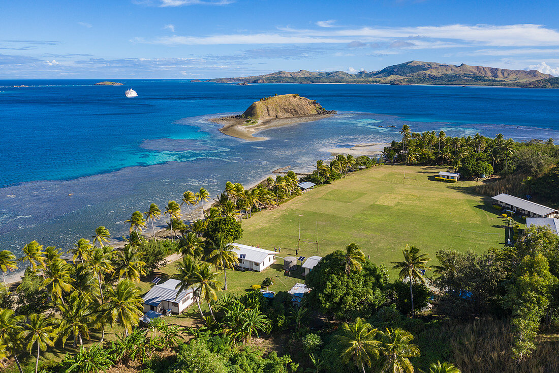 Luftaufnahme der Dorfschule mit Kreuzfahrtschiff MV Reef Endeavour (Captain Cook Cruises Fiji) in der Ferne, Nabukeru, Yasawa Island, Yasawa Group, Fidschi-Inseln, Südpazifik
