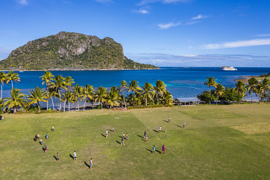 Luftaufnahme von Jungen die Rugby auf dem Feld der Dorfschule spielen mit Kreuzfahrtschiff MV Reef Endeavour (Captain Cook Cruises Fiji) in der Ferne, Nabukeru, Yasawa Island Yasawa Group, Fidschi-Inseln, Südpazifik