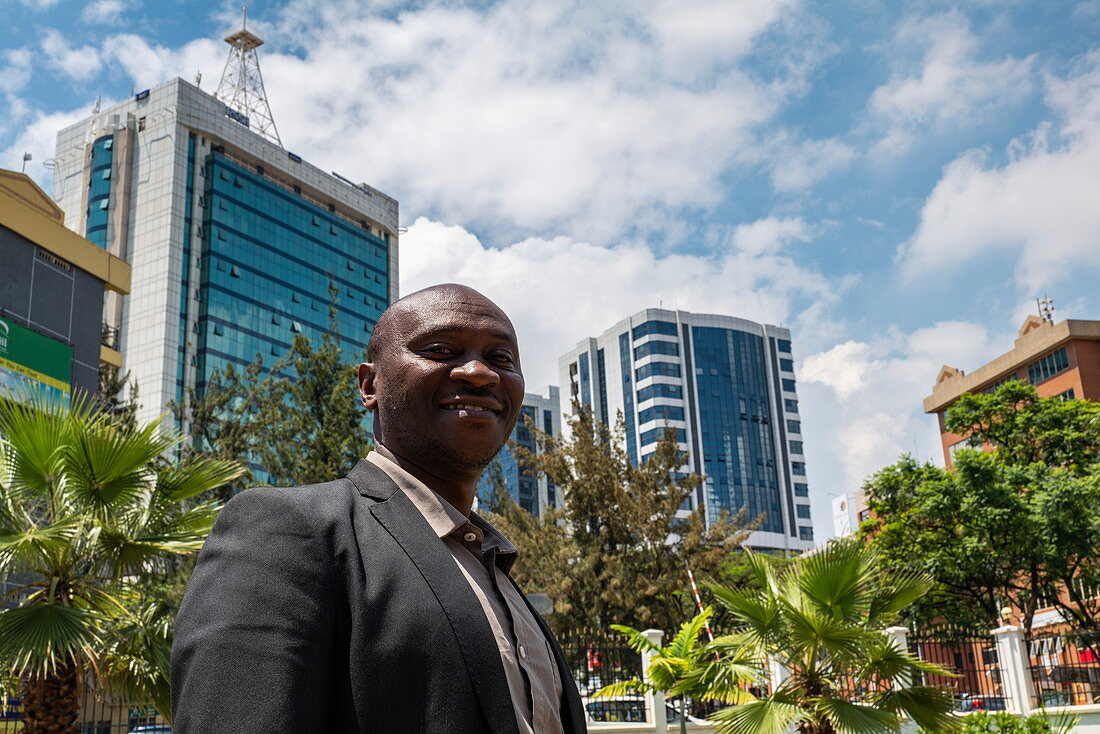 Würdevoll blickender ruandischer Mann posiert vor Parklandschaft und Bürohochhäuser in der Innenstadt, Kigali, Kigali Province, Ruanda, Afrika