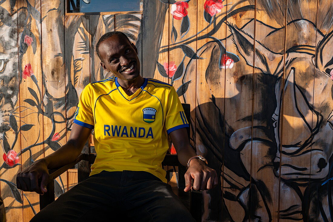 Young man with Rwanda jersey sits in front of mural with safari animal motif in a cafe, Kayonza, Eastern Province, Rwanda, Africa