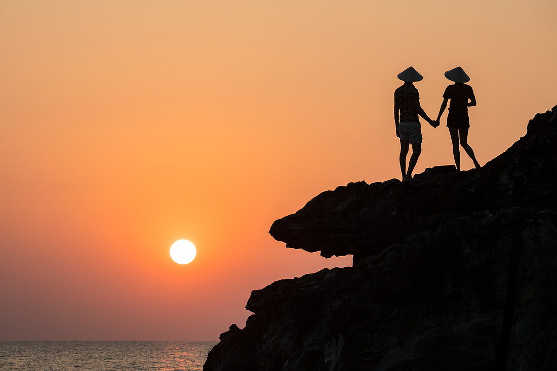Silhouette von jungem Paar das konische Hüte trägt und von Felsvorsprung neben Dinh Cao Schrein bei Sonnenuntergang auf Meer hinaus schaut, Duong Dong, Insel Phu Quoc, Kien Giang, Vietnam, Asien
