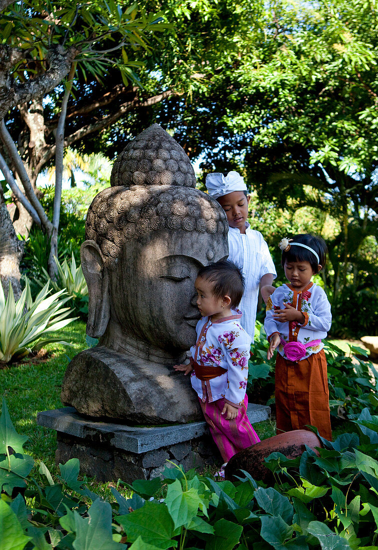 Drei balinesische Kinder spielen in traditioneller Kleidung um eine riesige Buddha-Statue in tropischer Umgebung Bali, Indonesien, Asien
