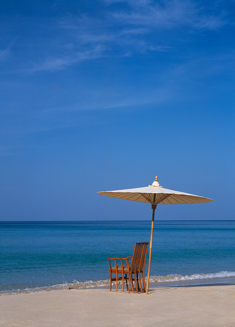 A rattan chair and an umrella, shot on a beach in Phuket, Thailand, against a clear blue ocean and sky.