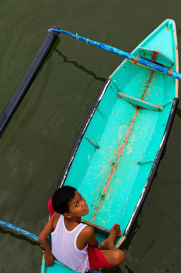 Ein junger malaysischer Junge, der von seinem türkisfarbenen Boot aus in die Kamera schaut, von oben in Borneo, Malaysia