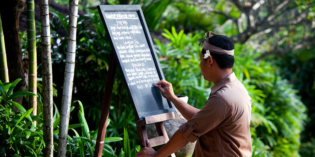 Ein balinesischer Kellner, der das Tagesmenü auf einer Tafel in einer tropischen Umgebung schreibt. Singaraja, Bali, Indonesien, Asien