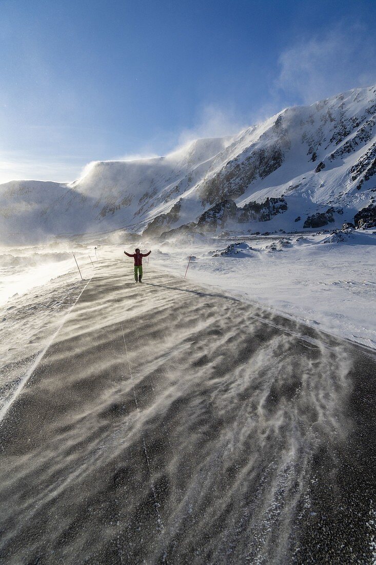 Man with arms raised standing in the windstorm on frozen coastal road, Barents Sea, Berlevag, Varanger Peninsula, Finnmark, Norway