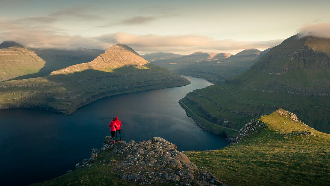 Couple at Hvithamar near the town of Gjogv on the Faroe island of Eysturoy with a panoramic view of the fjord at sunset