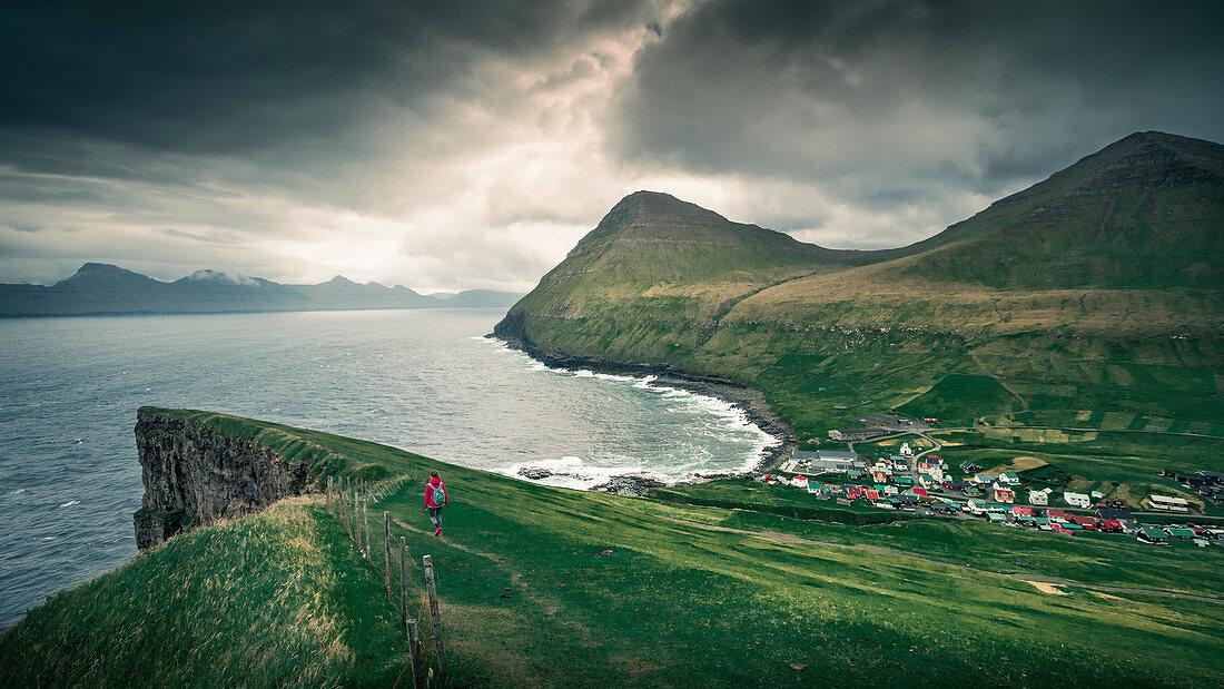 Woman hikes to village Gjogv on Eysteroy with gorge, sea and mountains, Faroe Islands