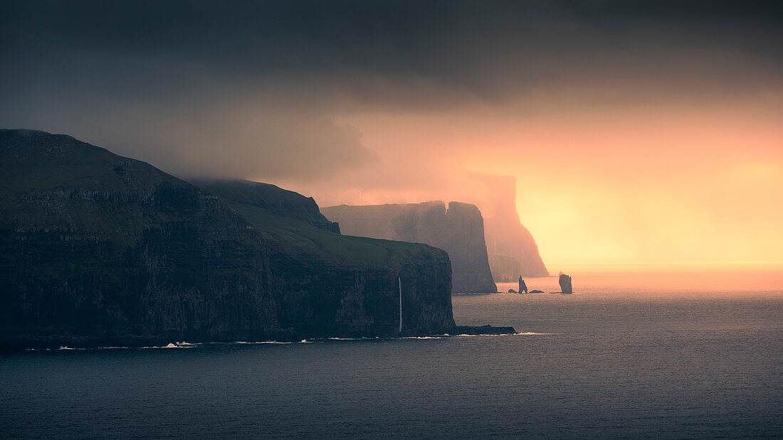 Sunset with clouds on the coast of Eysteroy with steep cliffs and rock formation Risin og Kellingin, Faroe Islands