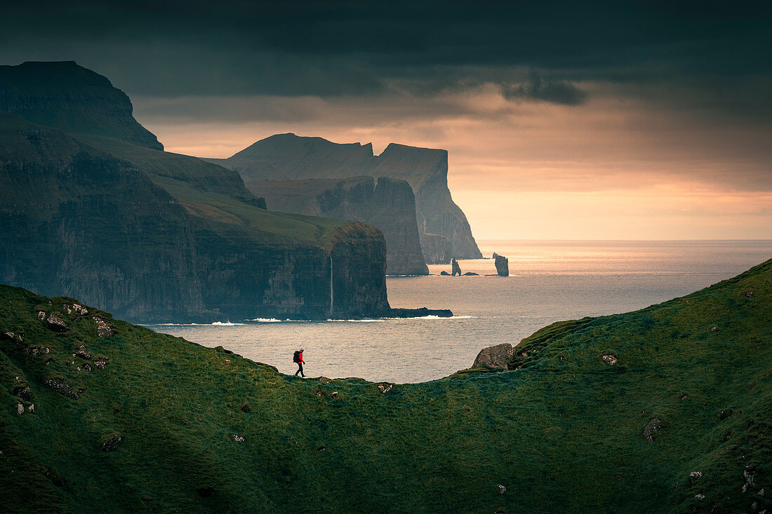 Mann wandert auf Klippe auf der Insel Kalsoy, Steilküste und Felsen von Eysteroy im Hintergrund im Sonnenuntergang, Färöer Inseln\n