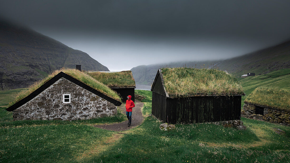 Man in red jacket in front of huts with grass roofs in the village of Saksun on Streymoy Island, Faroe Islands