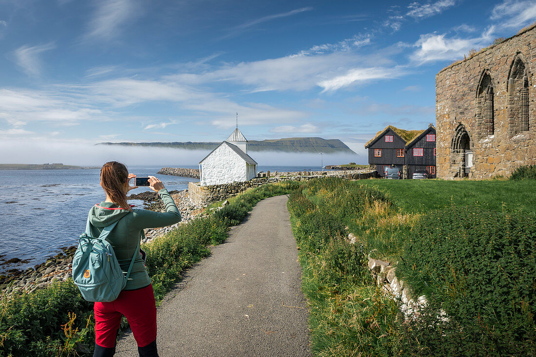 Woman photographs the church and ruin in the village of Kirkjubøur on Streymoy, Faroe Islands