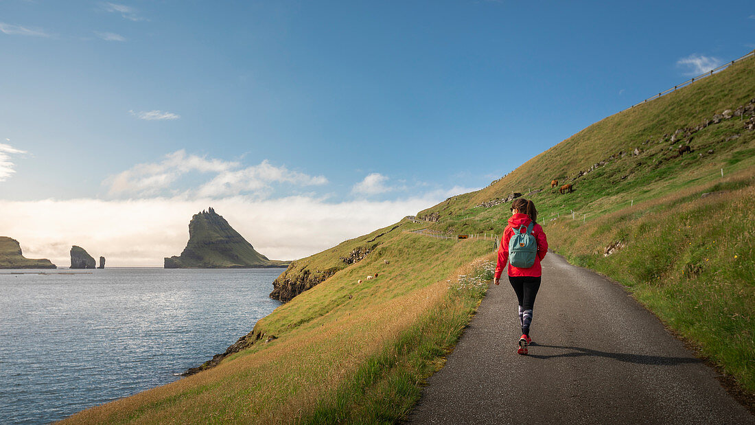 Woman hikes in sunshine in front of Drangarnier rock formations on Vagar, Bour, Faroe Islands