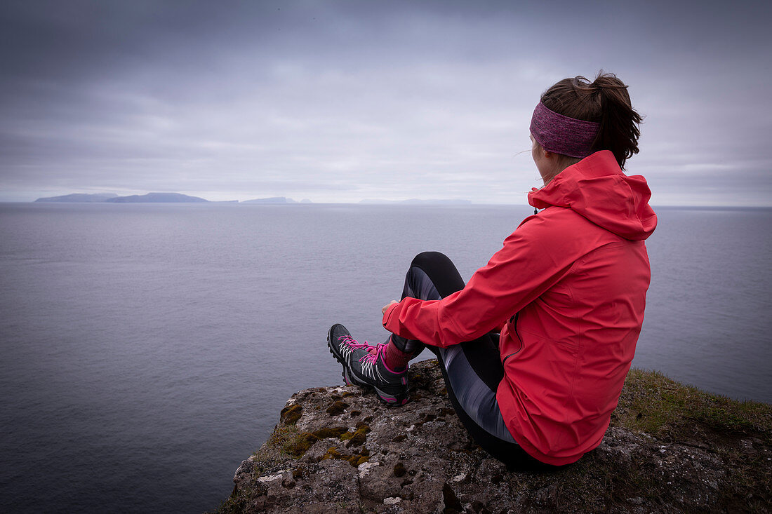 Frau mit pinker Outdoorjacke an der Klippe mit Blick auf das Meer, Färöer Inseln\n