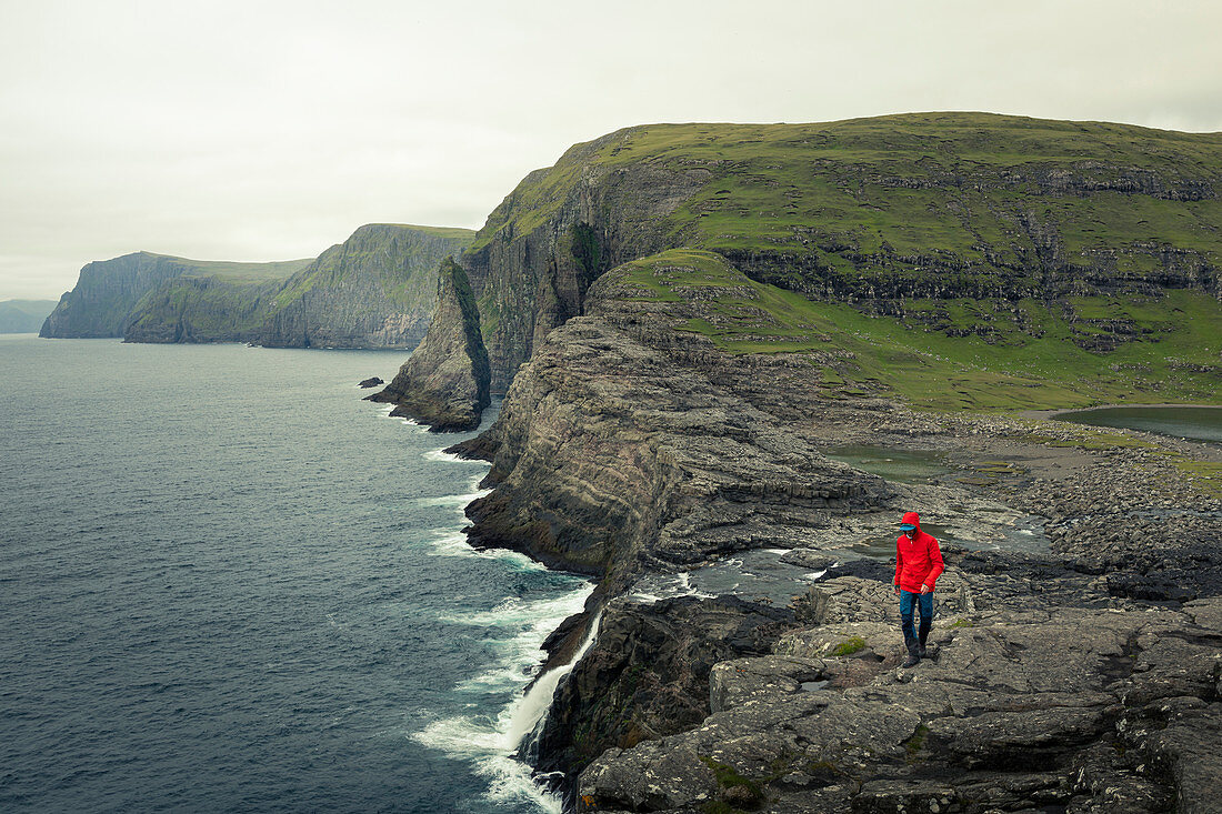 Man in red jacket on coast, waterfall and cliffs at Trælanípa on the island of Vagar, on Lake Leitisvatn, Faroe Islands