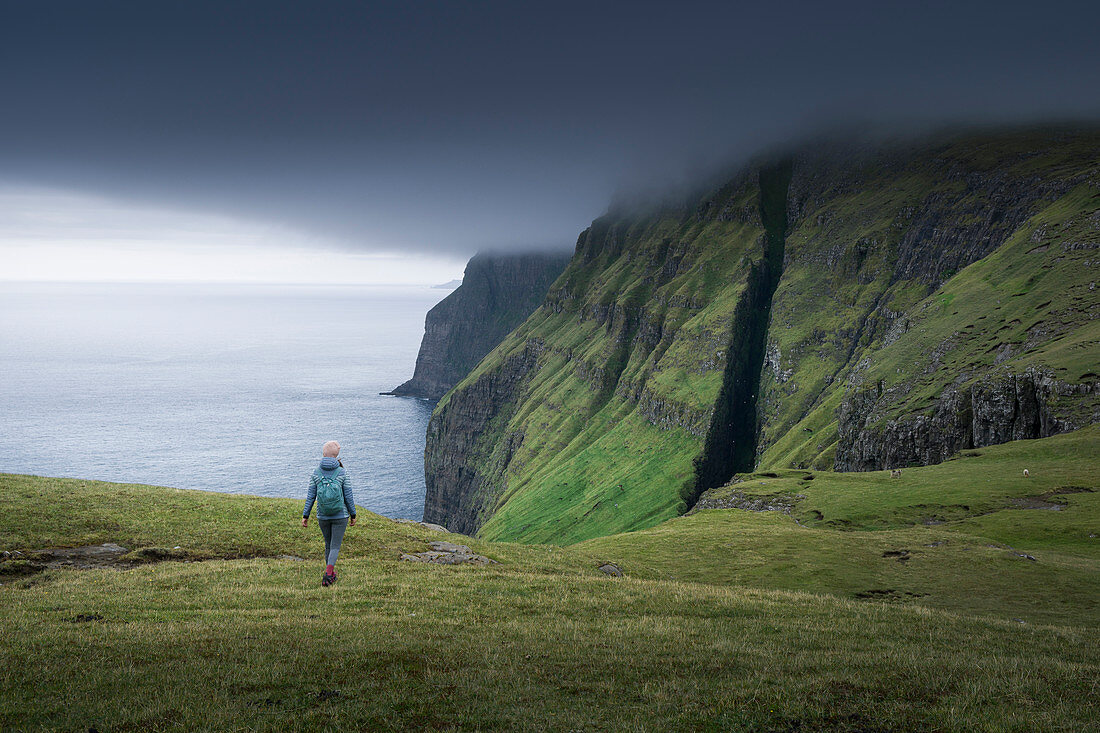 Frau wandert an der Steilküste von Dunnesdrangar unter dunklen Wolken auf Vagar, Färöer Inseln\n