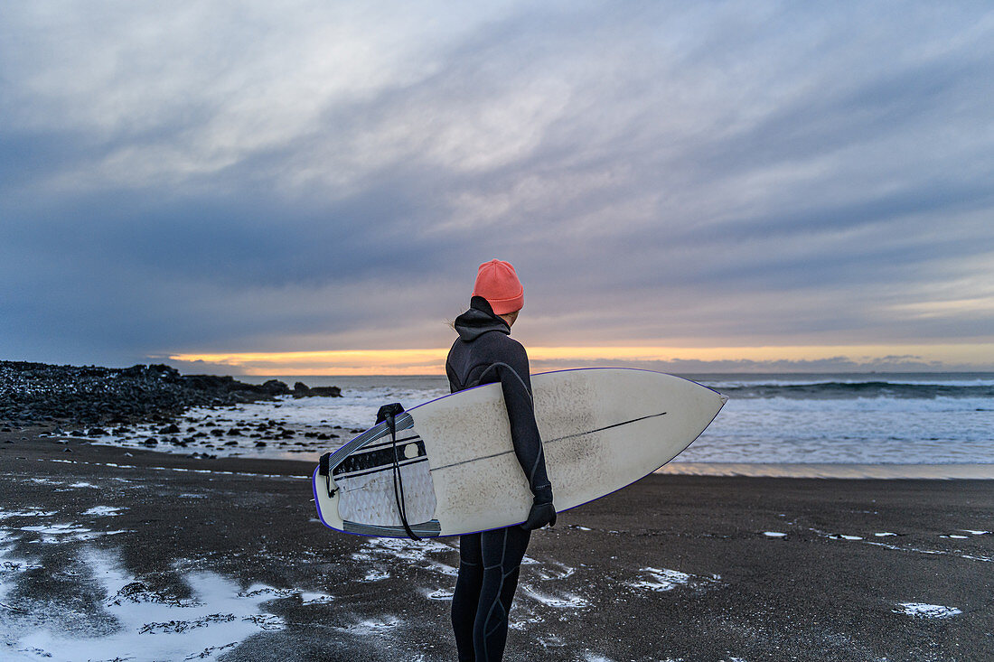 Frau in einem Neoprenanzug an einem Strand, der ein Surfbrett hält und auf Meer schaut.