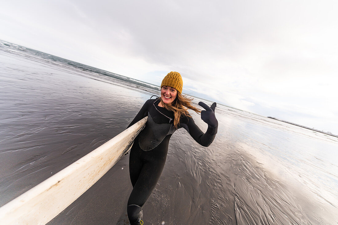 Eine Frau, die einen Neoprenanzug trägt und ein Surfbrett trägt, das über einen schwarzen Strand mit dem Meer im Hintergrund geht.