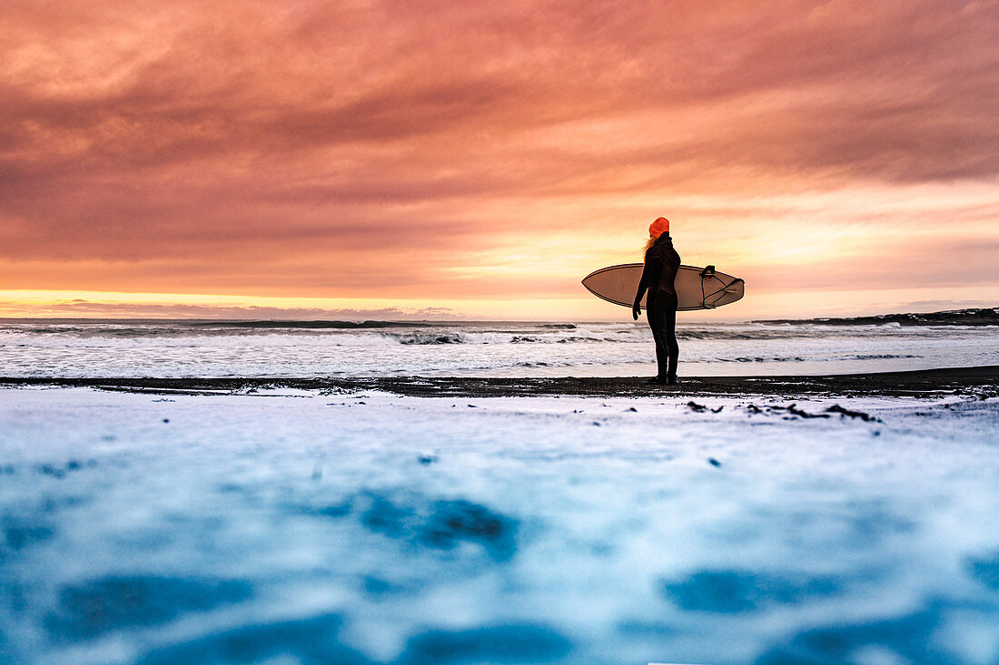 Rear view of  a woman standing on a beach holding a surfboard, looking out to sea with a sunset in the background.