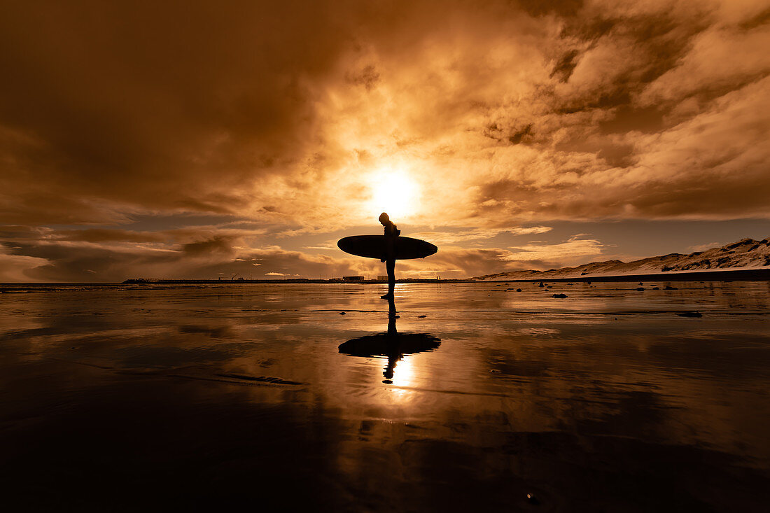 Schattenbild einer Frau, die ein Surfbrett trägt, das über einen Strand mit einem Sonnenuntergang im Hintergrund geht.