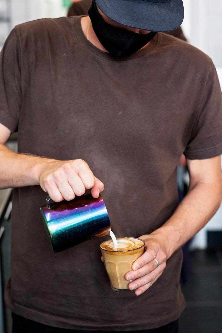 Close up of male barista wearing face mask pouring cafe latte.