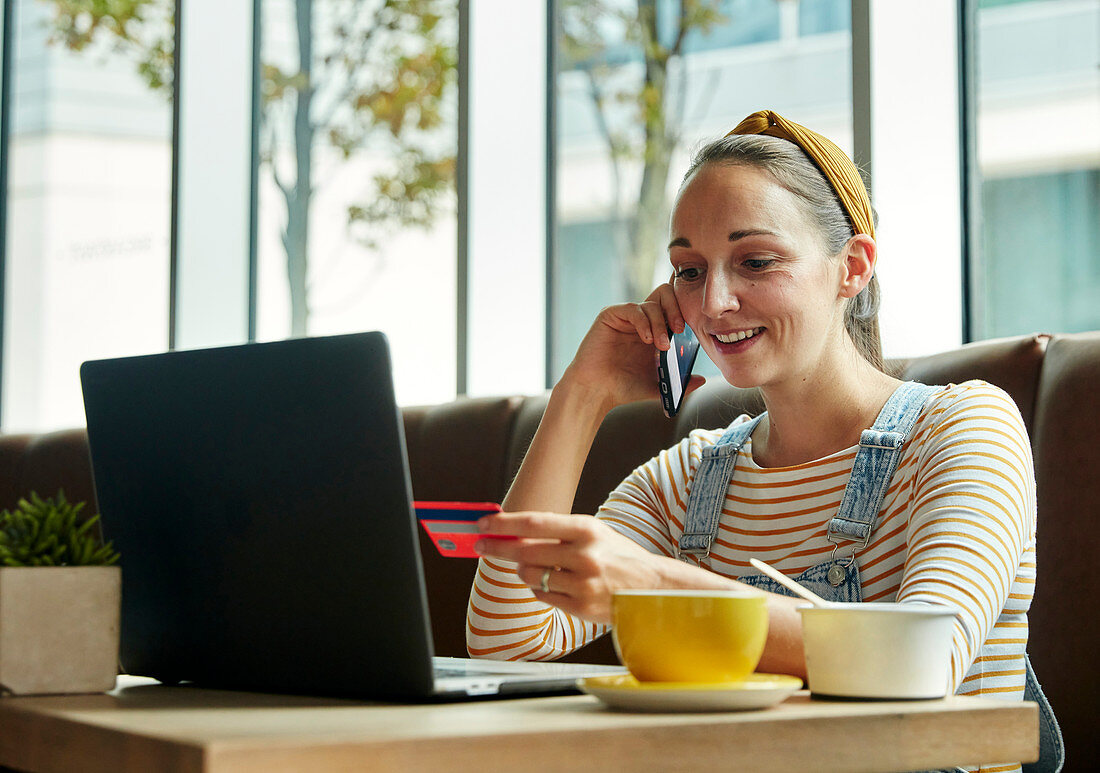 Frau sitzt in einem Café mit einem Laptop und telefoniert mit einem Smartphone, hält eine Kreditkarte