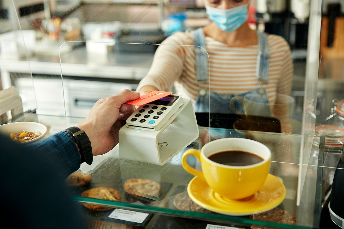 Woman in a face mask behind cafe counter with safety screen, offering a contactless payment terminal to a customer