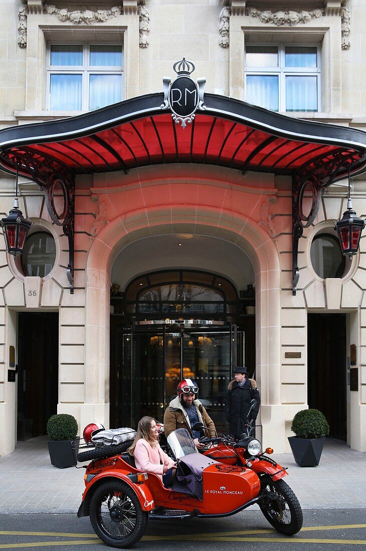 Frankreich, Paris, Royal Monceau Hotel, Frau, die in einem Retro-Seitenwagen vor der Hotelfassade reitet, bewacht durch zwei Kammerdiener